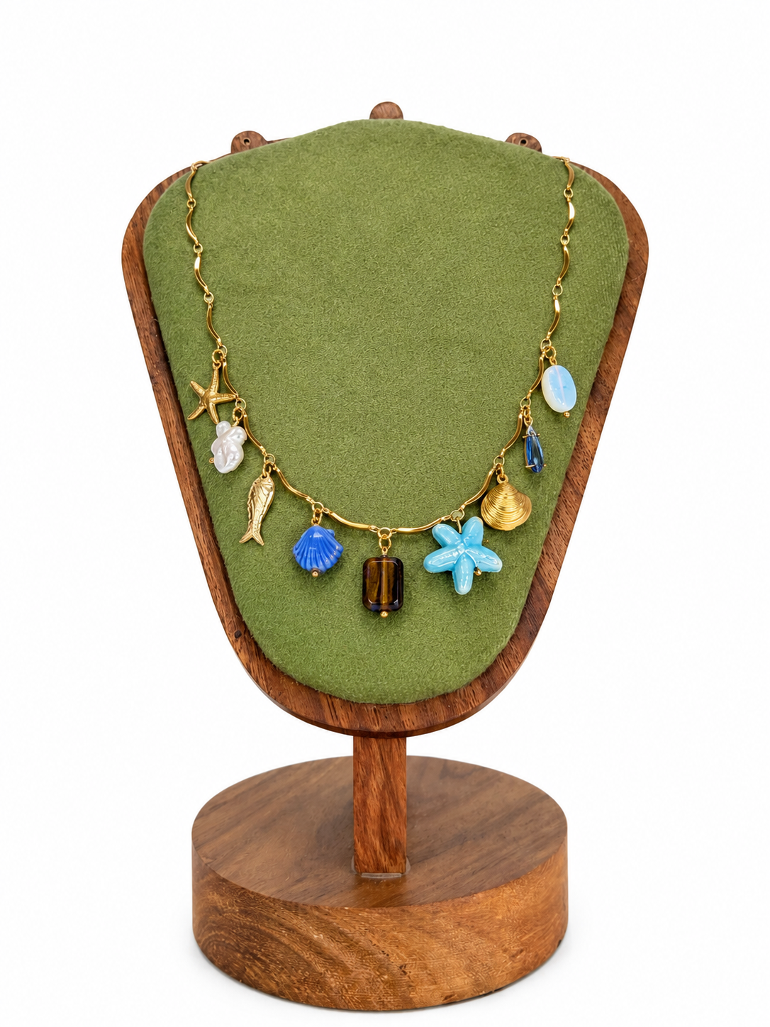 A gold charm necklace displayed on a green velvet jewelry bust with a wooden stand, featuring beach-themed charms including a starfish, seashell, fish, and blue starfish.